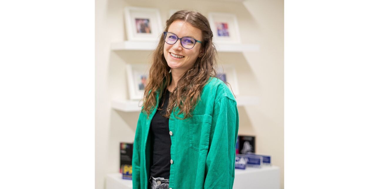 A portrait of Leah Elston-Thompson, a white woman with long brown hair wearing glasses and a green shirt over a black top stands in front of shelving